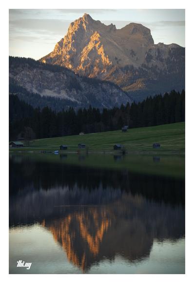 Photo of a mountain (Aggenstein) illuminated in orange light by the sunset/alpenglow, towering in the distance and reflecting in the completely still water surface of the mountain lake in the foreground. In between, near the lake, gentle rolling meadows with several small wooden huts then forests and deep valleys behind. Almost clear sky with some streaky clouds. A refuge (Bad Kissinger Hause) is visible on the ridge on the right side of the mountain.