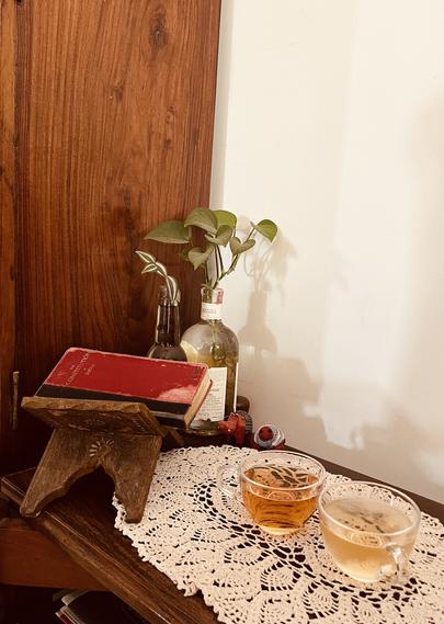 A cozy tabletop scene featuring two tea cups—one with amber tea and the other with clear tea—set on a lace doily. A red book titled "The Constitution" rests on a wooden book stand, alongside a couple of decorative bottles with water plants in them.