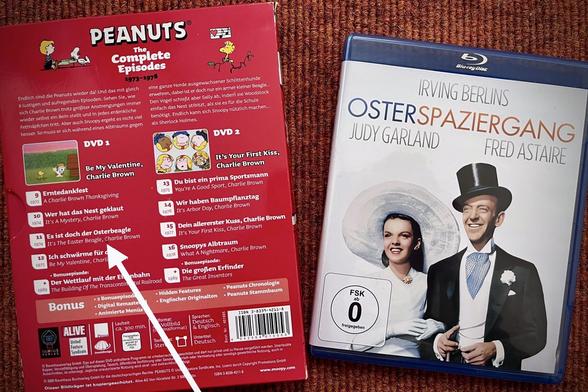 Eine DVD Box und eine Blu Ray Hülle sind zu sehen. Links die Rückseite einer DVD Box, die mehrere klassische Fernsehfilme der Comic Figuren „The Peanuts“ von Charles M Schulz enthält. Ein weißer nachträglich ins Bild einfügtet Pfeil zeigt auf die Episode: „Es ist doch der Osterbeagle“. Rechts ist dei BluRay Hülle des Films „Osterspaziergang“ zu sehen auf dem Judy Garland und Fred Astaire Arm in Arm und in festlicher Kleidung abgebildet sind. Auch der Name Irving Berlin wird erwähnt.
