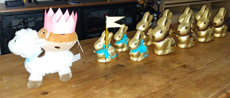 A (soft toy) hot cross bun is wearing a paper crown and riding on a lamb at the head of a Lindt bunny army. The first bunny carries a post-it and pencil banner.