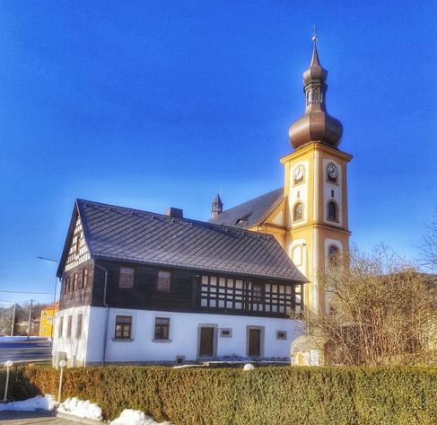 Kirche in der Gemeinde Skalná, Tschechische Republik. Blauer, wolkenloser Himmel. Im Vordergrund eine Hecke, dahinter ein noch kahler Baum.