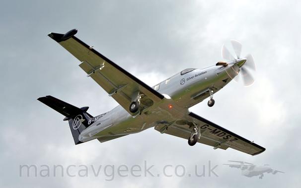 Low aside view of a silvery-grey, single engined BizProp flying from left to right at a very low altitude, with undercarriage lowered and flaps extended behind the wings, suggesting it is about to land.
The plane has black "Silver Aviation" titles on the forward fuselage, under the cockpit windows, and the registration "G-MDSZ" in black under the left wing.
The registration also appears in white on the black fillet attaching the tail to the upper fuselage.
The tail is also black, with the white outline of a circle containing an outline of a mountain in the middle.
A bright white light is shining forward from 2 thirds of the way along the right wing, with a red beacon also shining under the fuselage.
Grey sky fills the rest of the frame.