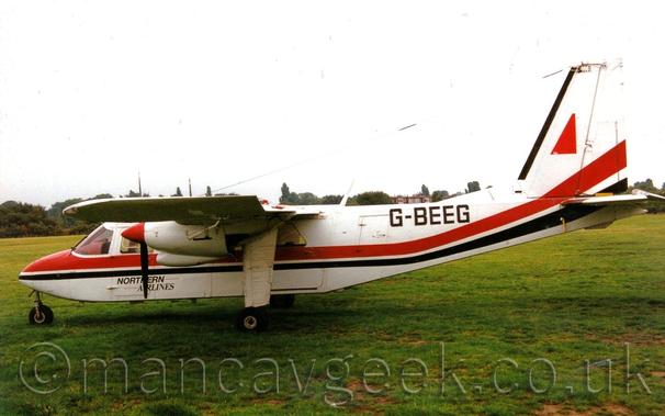 Side view of a small, boxy, white, high-winged, twin propellor-engined airliner parked on grass facing to the left.
The plane is mostly white, with a thick red and thin black stripe running along the body before swooping up into the lower reaches of the tail.
There are small, black "Northern Airlines" titles on the lower forward fuselage, and larger, black registration "G-BEEG" on the upper rear.
The landing gear is fixed, with a nose-wheel at the front, and the main wheels on long white struts that come down from the wings, just inboard of the engines.
Green grass fills the foreground, leading up to trees lining the airfield perimeter in the distance, under flat grey skies.