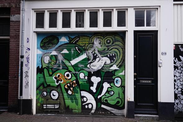 Façade d’immeuble avec une porte noire à droite. À gauche, street art composée de différents monstres verts grimaçants. Encadrement en blanc.