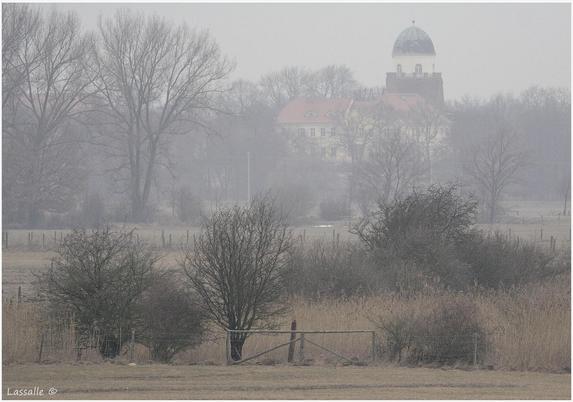 Die Burg Lenzen liegt im morgendlichen Dunst, inmitten noch laubloser Bäume. Gut zu sehen ist der große Turm der Burg, der mit einer halbkugelförmigen Kuppel bedeckt ist.
Mehrere kleine, eingezäunte Weiden werden durch Sträucher begrenzt, zwischen den großen Sträuchern Im Vordergrund findet sich ein Gatter als Zugang zu einer Weide.
Das Foto entstand von einem gegenüber der Landschaft erhöhten Hinterdeich.
