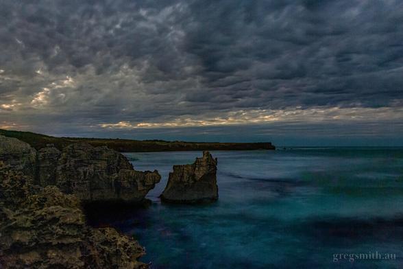 An overcast night sky, with some hints of moonlight poking through, and limestone cliffs meeting the ocean below.
