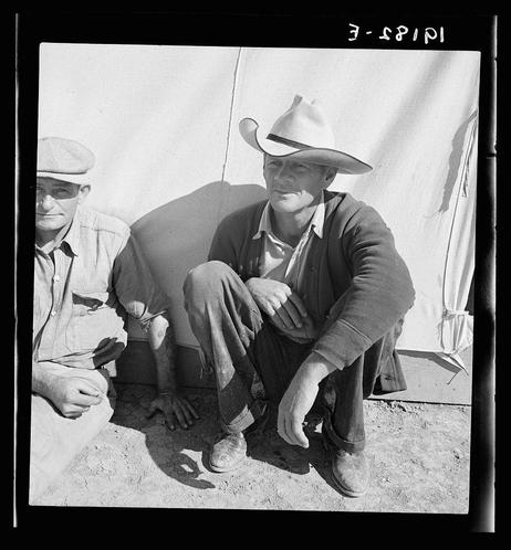 The photograph is a black and white image depicting two men seated on the ground, likely within an outdoor setting such as a camp. The man in the foreground wears a wide-brimmed hat with sunglasses atop it, indicating possible sun exposure or glare from sunlight. He dons casual attire consisting of layered shirts—denim over wool—and jeans, along with dark shoes showing signs of wear.

The seated men appear contemplative or possibly fatigued; their relaxed posture and downcast gazes suggest a momentary respite amidst daily laborious activities typical to migrant workers during the era. The setting's simplicity—the absence of luxury items—alongside their modest clothing points towards economic hardship, aligning with historical accounts of migratory work conditions.

The background is minimally detailed but includes what appears to be part of an awning or tent-like structure on right and a partial view of another individual in the far distance. The overall composition underscores themes prevalent during the Great Depression era concerning rural laborers' living circumstances, mobility, economic dependence, and social isolation within their transient community.

The image's historical context is further enriched by its accompanying information indicating that it was taken at "Migrant worker in camp" with a specified location of California—specifically identified as Yuma. The photograph captures the essence of migrant life d [...]