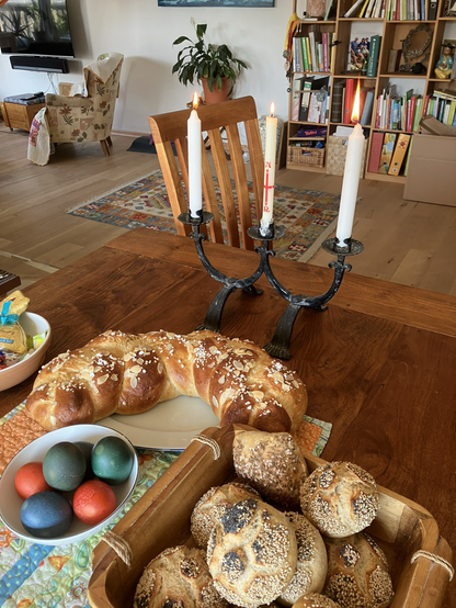 - *Kerzenständer*: Ein dreifacher Kerzenständer mit drei weißen Kerzen. - *Osterbrot*: Ein großes, ringförmiges Osterbrot mit Mandeln. - *Gebäck*: Eine Vielzahl von kleinen, gebackenen Brötchen in einem Holzkorb. - *Ostereier*: Ein paar bemalte Ostereier in einem Schälchen. - *Tisch*: Ein brauner Esstisch. - *Sessel*: Ein paar Sessel im Hintergrund. - *Wohnzimmer*: Ein gemütliches Wohnzimmer mit einem Fernseher und einem Bücherregal im Hintergrund