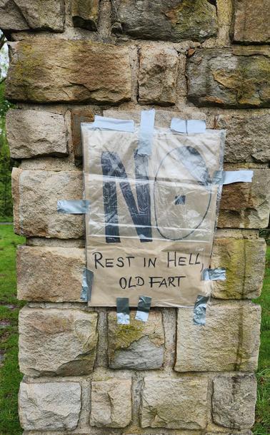 An einer schmalen Stele aus gemauerten Sandsteinquadern hängt ein mit schwarzem Filzstift selbst gemaltes  und geschriebenes Plakat mit den Worten no in Großbuchstaben. Das o ist ein mit wenigen Strichen gezeichnetes Hitler-Gesicht mit Scheitel und Popelbremse. 
Unter dem no steht Rest in Hell, old fart.
Das Plakat ist in Plastik gehüllt gegen Regen.
