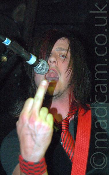 Close-up of the head and upper body of a man with long dark hair covering his right eye, wearing a grey t-shirt, a black waistcoat, and a red tie with thin black stripes, holding up an extended middle finger on his right hand to the camera, with his mouth open, about to stick his tongue out.
A red guitar strap is slung over his left shoulder, and there is a microphone on a stand by his mouth on the left of the frame.
Black walls and ceiling fill the rest of the frame.