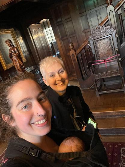 A selfie of three people in an astonishingly wood panelled room at a historic property in England (with hints of decadent no-sitting-here chairs, wooden sculptures, and big paintings). At the back is an older white woman with short cropped white hair, black clothes, and a big smile. In the foreground is a smiling white woman with dark hair pulled back and a baby carrier, in which we can just about see the top of a baby's head.