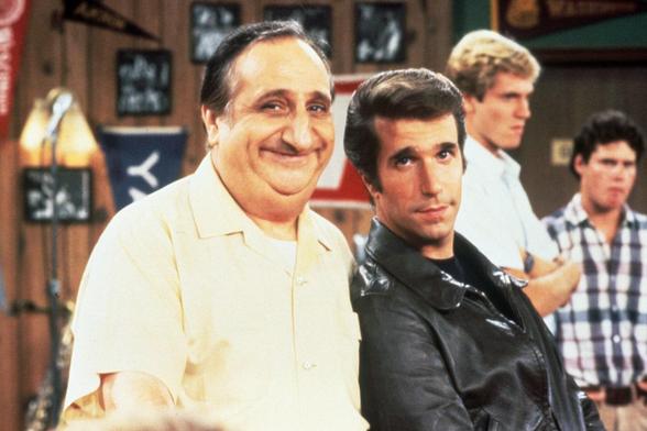 A photo from the set of 1970s TV show "Happy Days". It's "Big Al's" diner. In centre is Big Al and "The Fonze", played by Al Molinaro and Henry Winkler respectively. Al is smiling and is a dark haired balding largish Italian-American man in a pale short sleeved shirt. The Fonze is The Fonze in a black leather jacket, looking cool.