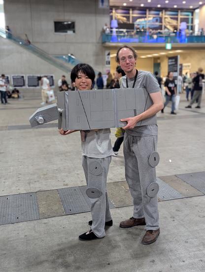 A couple dressed in grey—with their legs as the the legs of an AT-AT—and a painted cardboard head and body encompassing both of them.