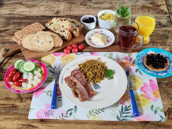 Plank met knäckebröd, paasstol en afbak broodjes. Klein schaaltje met blauwe bessen jam. Iets groter schaaltje met kip kerrie salade, longdrink glas met mock mojito en takje verse munt, glas sinaasappelsap, glas thee, schoteltje met pannenkoekje met blauwe bes, schoteltje met stukje boter, ontbijtbordje met bacon en tofu scrambled, schoteltje met snoeptomaatjes, plakjes komkommer en zilveruitjes en 5 pure chocolade paaseitjes.