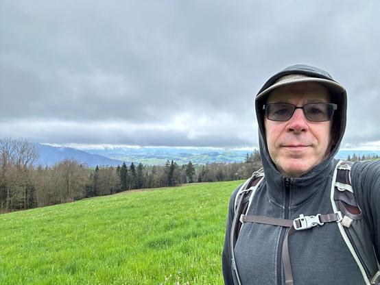 Selfie of me, a middle-aged white guy in sunglasses, a dark gray hoodie with the hood up and wearing an olive baseball cap. I’m standing in front of a green pasture under an overcast sky with some trees and a valley visible in the distance.