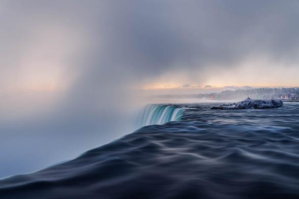 View from the edge of Horseshoe Falls at Niagara, where the water plunges into the mist on a cold winter morning with a soft sunrise glow on the horizon.