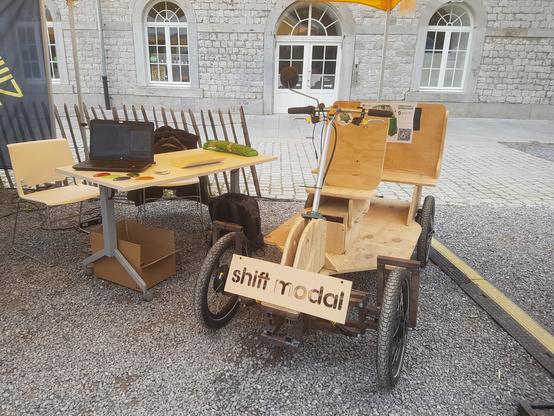 Un véhicule à 4 roues de vélo avec un plancher et sièges en bois, sous une tonelle à côté d'une table avec un ordi et des échantillons de plastique biosourcé. Sur l'avant du véli une plaque en bois avec "shiftmodal" découpé