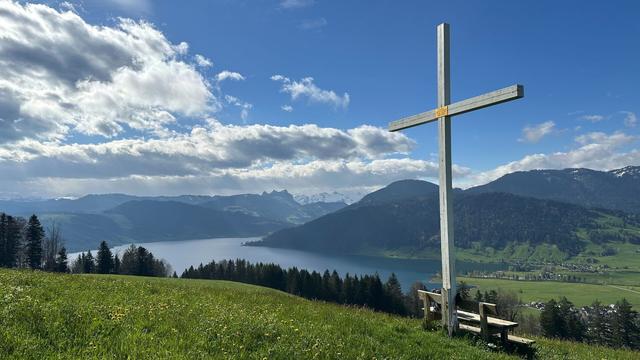 View of lake Ägeri under a partly cloudy sky. A white cross over a park bench, green pasture and trees are in the foreground while forested hills and distant mountain peaks are in the background.