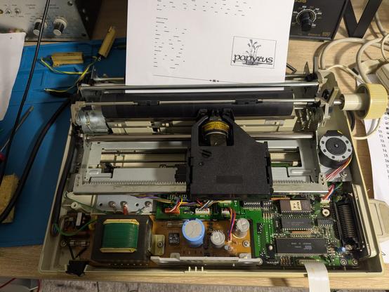 Une imprimante matricielle Star LC-10 sans son capot, on voit l'alimentation et le PCB principal,  ainsi que le chariot horizontal avec la cartouche contenant le ruban encreur. Il y a une feuille A4 imprimée avec un dessin d'une feuille de papyrus et le mot Papyrus en dessous