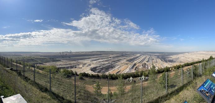 Das Bild zeigt den imposanten Blick vom Aussichtspunkt am Tagebau Hambach. Die riesige Grube erstreckt sich über die gesamte Bildbreite und ist geprägt von tiefen Abbau-Stufen und breiten Fördertrassen. Im Hintergrund sind gewaltige Schaufelradbagger zu erkennen. Im Vordergrund verläuft ein Sicherheitszaun, begleitet von junger Vegetation und mehreren Informationstafeln. Der Himmel ist klar mit einzelnen Wolken, was die Weite und Dimension der Landschaft zusätzlich betont.