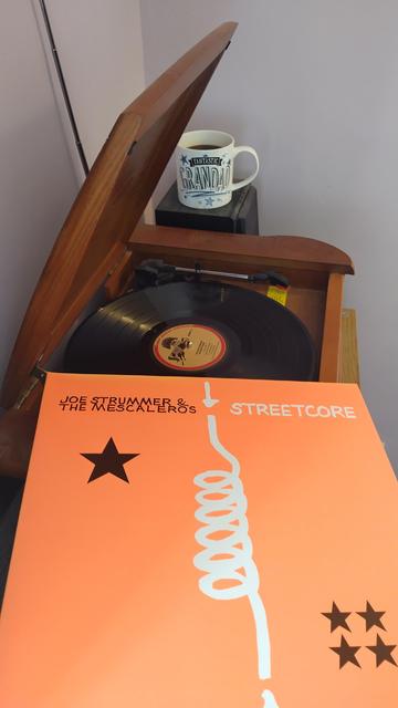 Record player with a 12" black vinyl record.
In the foreground is an orange album cover.
Behind the player is a mug of coffee on a speaker.