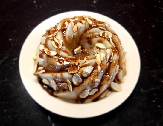 A swirled vegan almond pudding topped with slivered almonds and drizzled with a glossy caramel syrup, served on a white plate over a dark surface.