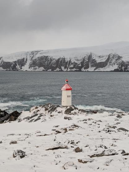 Small lighthouse surrounded by snow near a fjord. Grey sky.