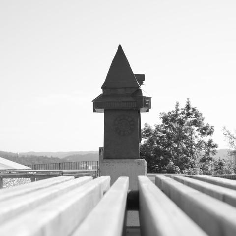A small interpretation of the clocktower in Graz. In the foreground are the wooden beams of a bench that lead exactly in the direction of the clock tower. The image is in black and white.