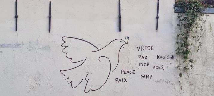 The intricate, stylised, black lined outline of a flying dove holding a tiny olive branch in its beak next to the word peace in different languages on a white, outside wall. A green leaf plant spills over the lower section of the wall next to the piece.