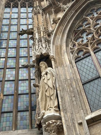 detail of a gothic cathedral: a robed, bearded statue on a podium in an alcove. Above it, an ornate column extends upwards, spouting a gargoyle. Behind, a grid of grids of rectangular glass panels, each of slightly different hue, extends upwards seemingly forever. An ornate gothic arch window is partly visible in the foreground