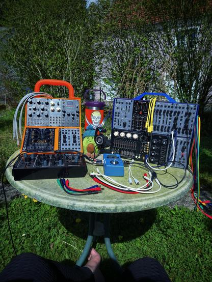 A starbase deep in Federation territory… captain’s log, stardate unknown. Two boldly colored, 3D-printed Eurorack cases (radiant orange and stellar blue) rest on a green, weathered garden table. Surrounded by blooming spring flora, the setup is ready for sonic warp trials. An orange thermos bottle adorned with a skull in Nonbinary pride colors stands tall beside the modules… radiating hydration and identity. The loyal officer AvocaJoe (knitted beanie included) guards the bridge. Multicolored patch cables arc like EPS conduits… connecting systems including a Stylophone drone synth below. Bare feet at the base… clearly, the Captain is grounded. Test phase initiated… tweaks may follow.