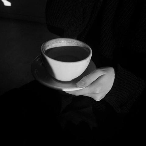 Eine Hand hält eine weiße Tasse mit Kaffee auf einem Unterteller, vor einem dunklen Hintergrund.