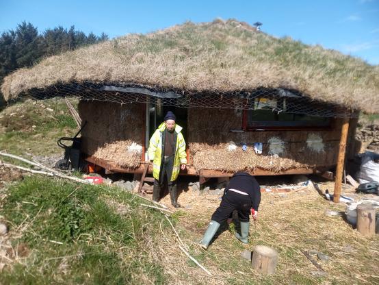 Outside the roundhouse. The shaggy turf roof hangs out far over the walls, to protect them from driving rain. The whole house appears to float a little off the ground. There are some trial patches of lime render.