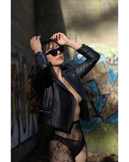 Eine Frau steht vor einer mit buntem Graffiti besprühten Betonwand, trägt eine Lederjacke und eine Sonnenbrille. Sie ist nicht in Pose, sondern wirkt, als würde sie sich gerade auf das Shooting vorbereiten. Der Moment wirkt ehrlich und ungestellt – ein spontanes Porträt mit urbanem Hintergrund.