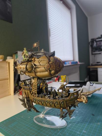 A miniature of a wooden skyship in a kind of steampunk aesthetic. Blades and metal reinforcements attached to the side, a big balloon at the top. Cannons are all along the railing and a giant one poking through the mouth of a skull at the front of a ship, posing as a figurine.
Four characters stand aboard. One Human-Albatros Hybrid has a sword raised, a small unpainted druid figure, a gold-iron painted Automaton with a giant shield and a psychic blade rogue on top of the balloon.