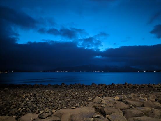 A rocky beach in the foreground with some stacked stones visible along the edge of a calm body of water. There is land across the water with lights along the edge and hills behind them. It is pre-dawn. Light is beginning to show behind clouds in the dark blue sky.