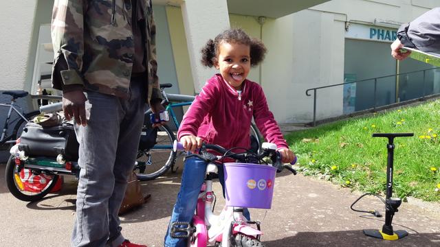 Fillette souriant de bonheur de pouvoir remonter sur son vélo tout juste réparé avec son père. Avec une carriole, 2 vélos, une pompe à pied et une pharmacie en arrière plan.