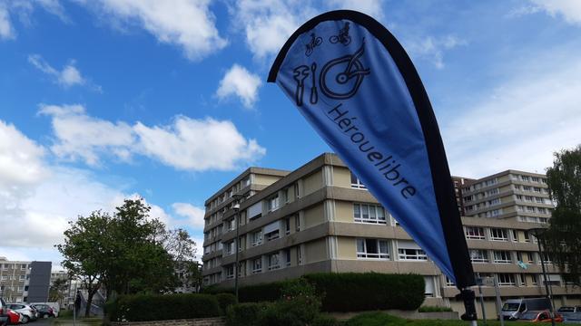 Oriflamme Hérouelibre avec des outils et l'arrière d'un vélo dessus entre ciel bleu + coton blanc, immeubles et arbre.