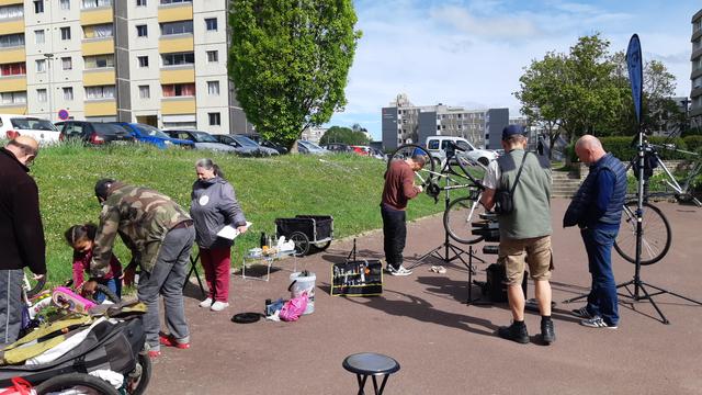 Atelier d'aide à la réparation de vélos sur une place piétonne près d'immeubles : Fillette et son père finissant de remonter un vélo enfant sous les yeux d'un et d'une bénévole de l'association ; homme nettoyant la transmission de son VTT sur un pied vélo ; hommes échangeant tout en vérifiant une roue de 700. Avec des carioles vélo, un panneau et une servante à outils ainsi qu'un seau avec brosses de nettoyage et un tabouret.