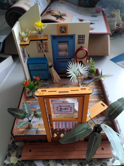 début de maquette d'une maison de jardin avec une porte en bois, une petite commode et diverses plantes en plastique ou en papier