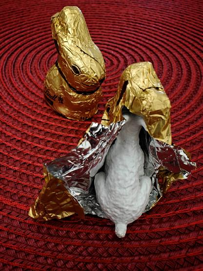 Chocolate Easter bunnies wrapped in gold foil on a red circular placemat. One bunny has been partially unwrapped, revealing a small white figurine of a dog that wears the foil like a disguise.