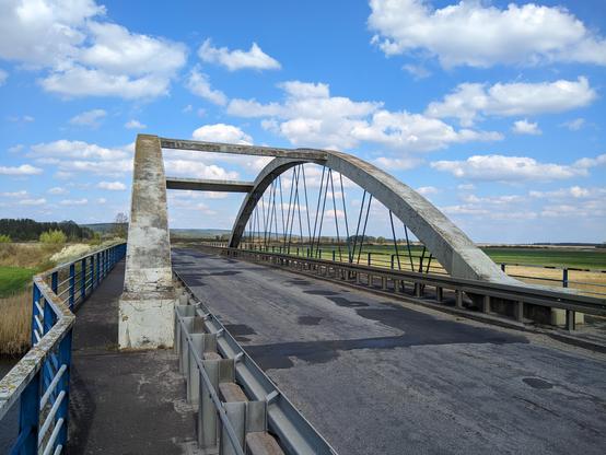 Most nad rzeką. Dwa betonowe łuki połączone poprzeczkami przechodzącymi nad szosą o zniszczonym asfalcie.