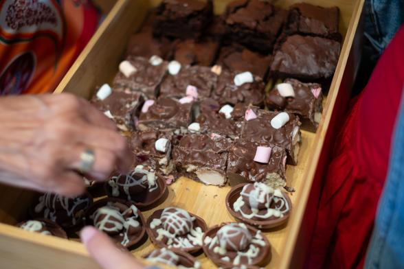 A wooden tray filled with various chocolate desserts, including brownies, rocky road squares with marshmallows, and round chocolates with white drizzled decoration. A hand is reaching for one of the desserts.