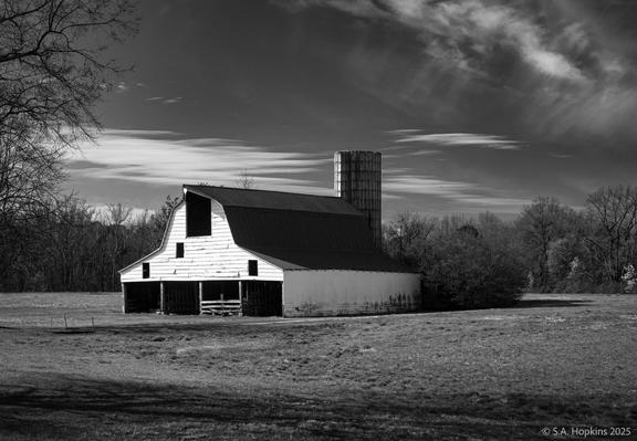 Black and white photograph of farmland featuring a barn reflecting the morning light.