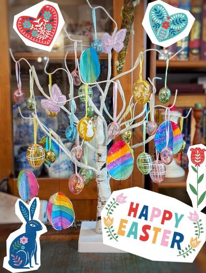 Kleines, birkenartig wirkendes Osterbäumchen, an den Ästen aus Draht hängen durchsichtige, gläsern anmutende, bunte Ostereier in klein und groß und papierne, vierseitige Ostereier, bemalt mit Wasserfarben. Vor allem die regenbogenfarbenen Seiten sind zu sehen. Im Hintergrund sind verschwimmen eine Vitrine mit Geschirr und ein Regal mit Büchern zu sehen. Sticker, die aussehen, wie aus Papier ausgeschnittene, osteuropäisch wirkende Ostermostive (Osterhase, Spruch "Happy Easter", Blüten, Herzen) rahmen das Bild.