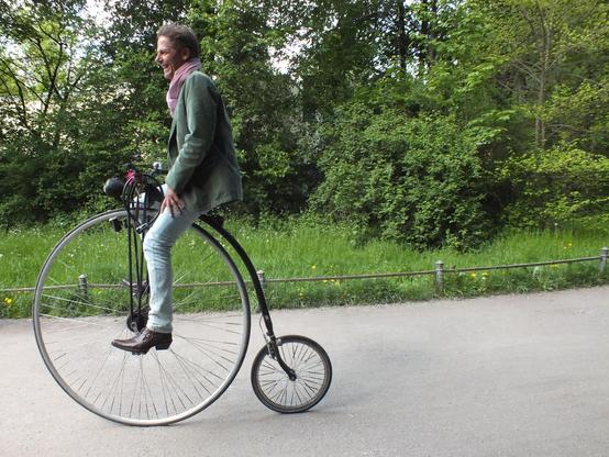 Person on a penny farthing on a paved path with grass and greenery behind. 
Pic near the Englischer Garten in Munich; 2014