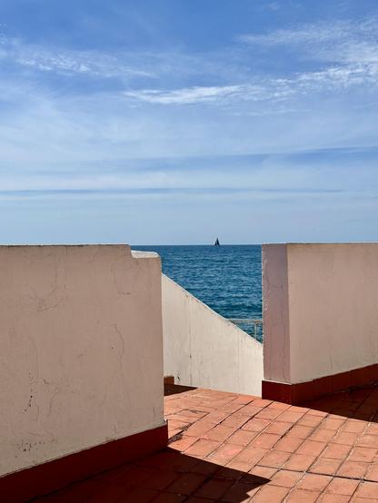 A seaside scene. The view from a terrace with white stucco walls and terracotta-red tiles in the foreground. The sun casts sharp, dark shadows across the floor, creating almost geometric surfaces of red, white, and blue. In the middle of the wall, there is an opening with a staircase leading down toward the sea. On the blue  horizon, a small dark sailboat is visible. The line of the sea horizon aligns perfectly with the top of the wall. The sky is mostly clear, with scattered clouds.

Pogled s terase z belim zidom in terakoto po tleh v ospredju na morsko gladino in nebo zadaj. Sonce meče ostre in temne sence čez tla. Skoraj geometrične ploskve rdeče, bele in modre. Na sredi zida je odprtina s stopniščem, ki vodi proti morju. Na modrem morskem horizontu je majhna temna jadrnica. Nebo je večinoma jasno, z nekaj koprenastih oblakov. Linija morskega obzorja je popolnoma poravnana z vrhom zidu terase.