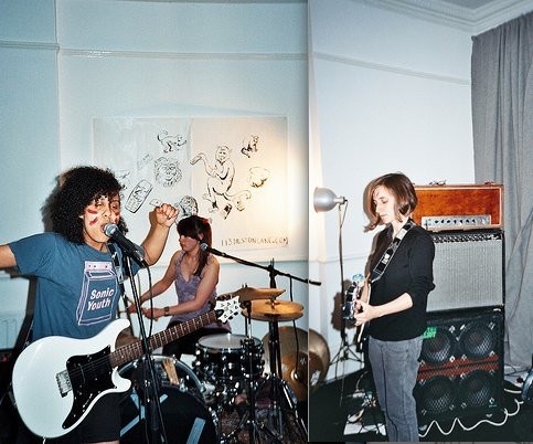 Trash Kit, a women & queer led post-punk band from early 2010s London, performing in a small venue. One member (Rachel Aggs) plays guitar and sings, another plays bass, while another is on drums. A poster with artwork is visible in the background, along with amplifiers and other music equipment.