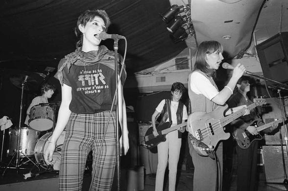 A black-and-white photo of early 80s female-fronted post-punk band Delta 5 performing on stage. The lead singer is in the foreground with a microphone, wearing a graphic t-shirt and plaid pants. Band members can be seen playing drums and electric guitars in the background.