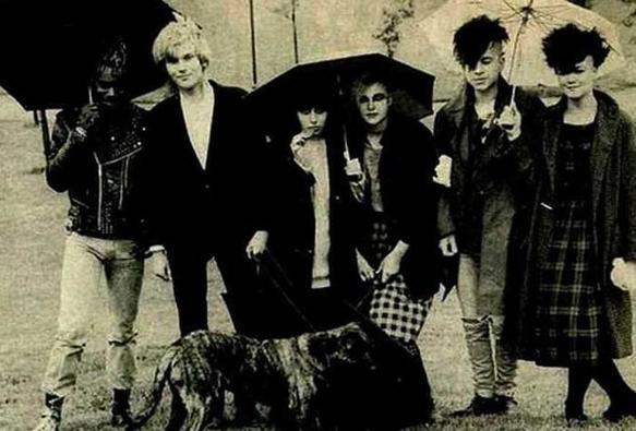 Black and white photo of 1980s London multi-gender, multi-racial anarcho punk band Hagar the Womb. All six members pose holding umbrellas in the rain with a dog in front of them.
